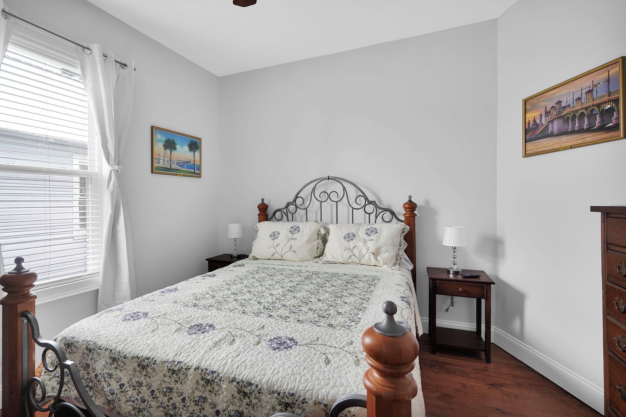 Een slaapkamer met een houten bed, nachtkastje, dressoir en een schilderij aan de muur.