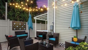 Un patio exterior con mobiliario de mimbre, un jacuzzi, luces colgantes y una piña decorativa.