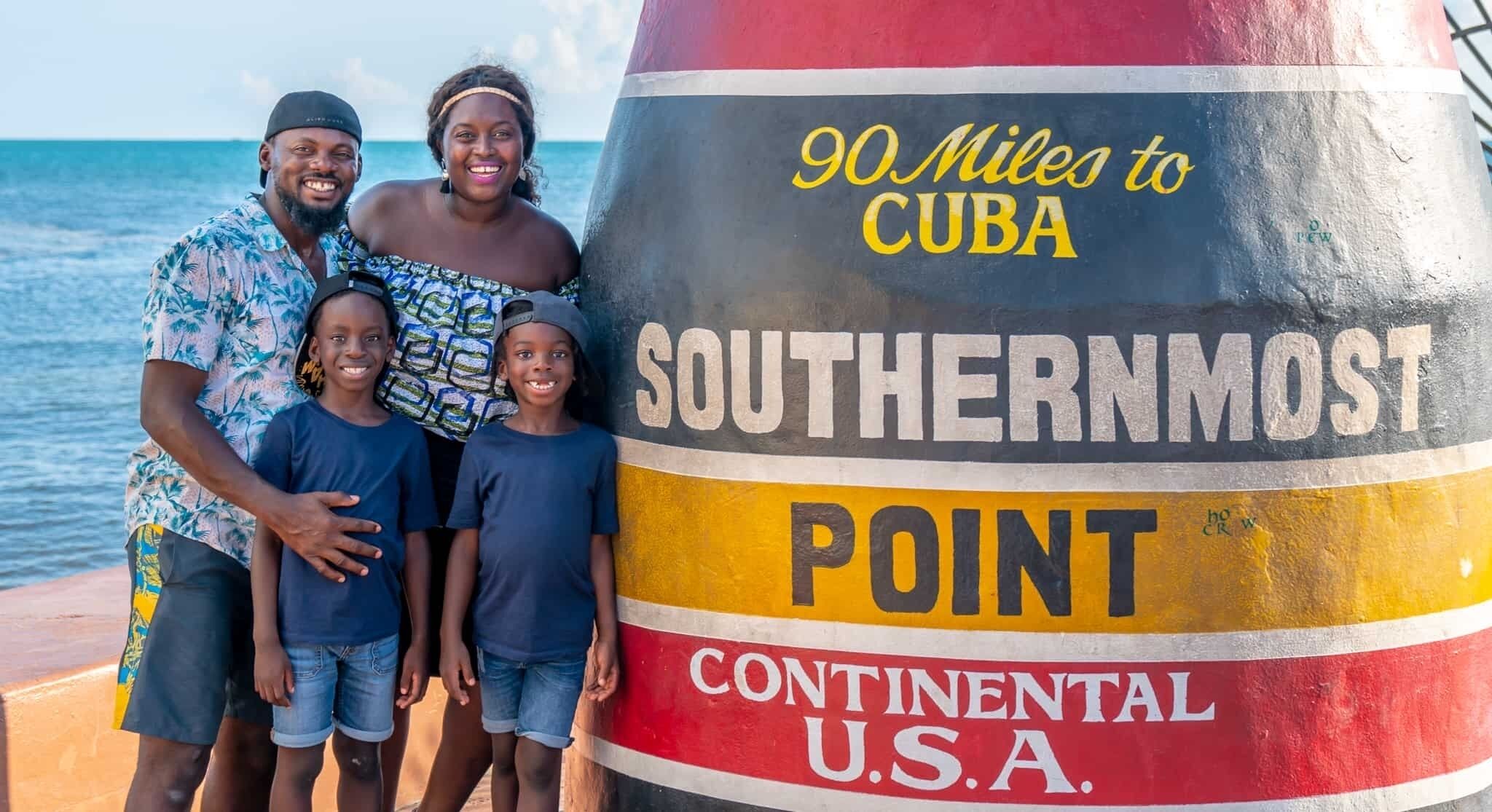 A family standing in front of a sign that reads "90 Miles to Cuba" and "Southernmost Point, Continental U.S.A."