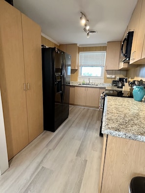 Una cocina con armarios de madera, un refrigerador negro, una encimera de granito y una ventana con persianas.