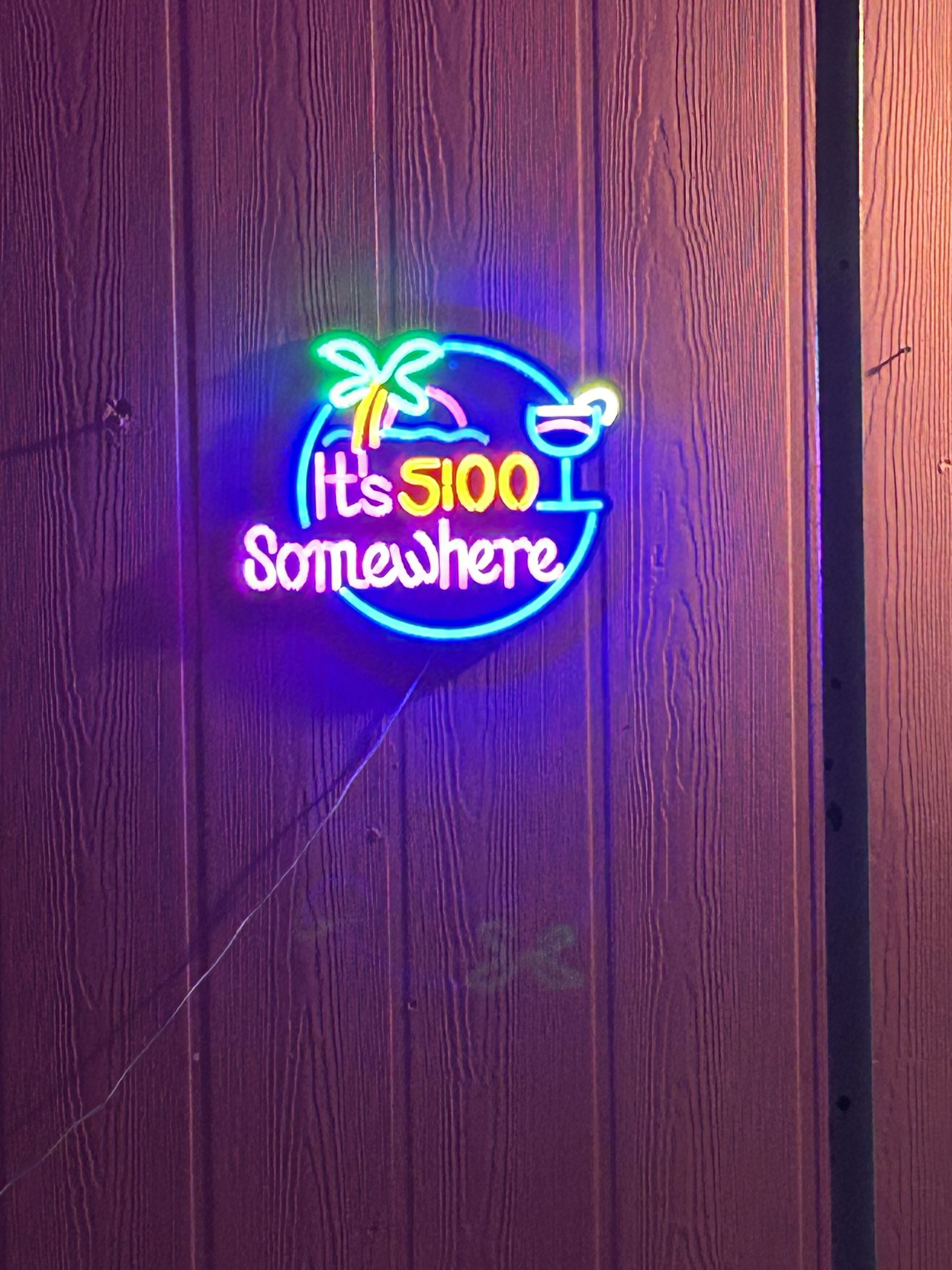 Ein leuchtendes Neon-Schild an einer Holzwand mit dem Schriftzug "It's 5100 Somewhere" sowie einem Palmen- und Cocktailglas-Symbol.