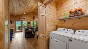 Interior de una cabaña de madera con zona de lavandería, un sofá y vistas a una terraza exterior por una puerta.