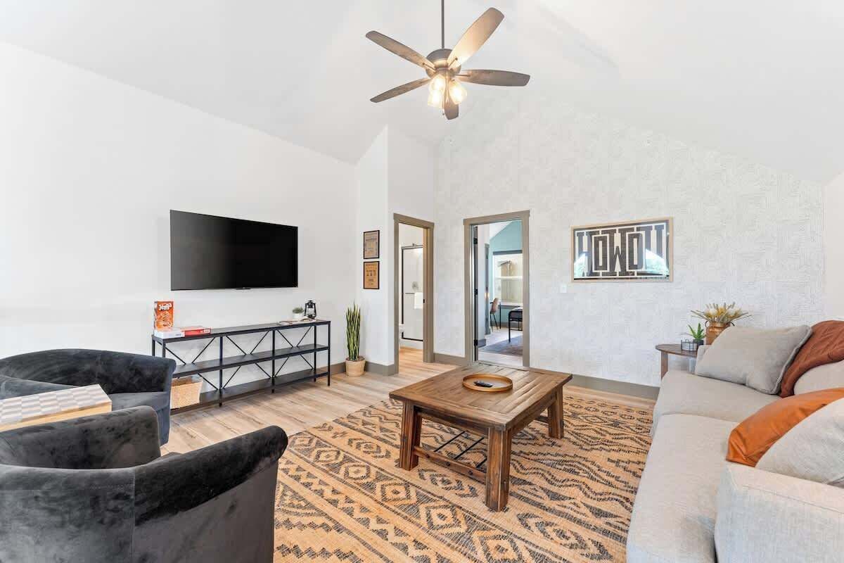 A modern living room with a flat-screen TV, a ceiling fan, a patterned rug, and a sofa.