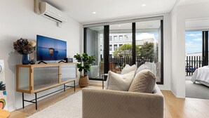 A modern living room with a large flat-screen TV, a beige sofa, a wooden cabinet, and a large window offering a view of the outside. - Creative and Carefree Living (Auckland)