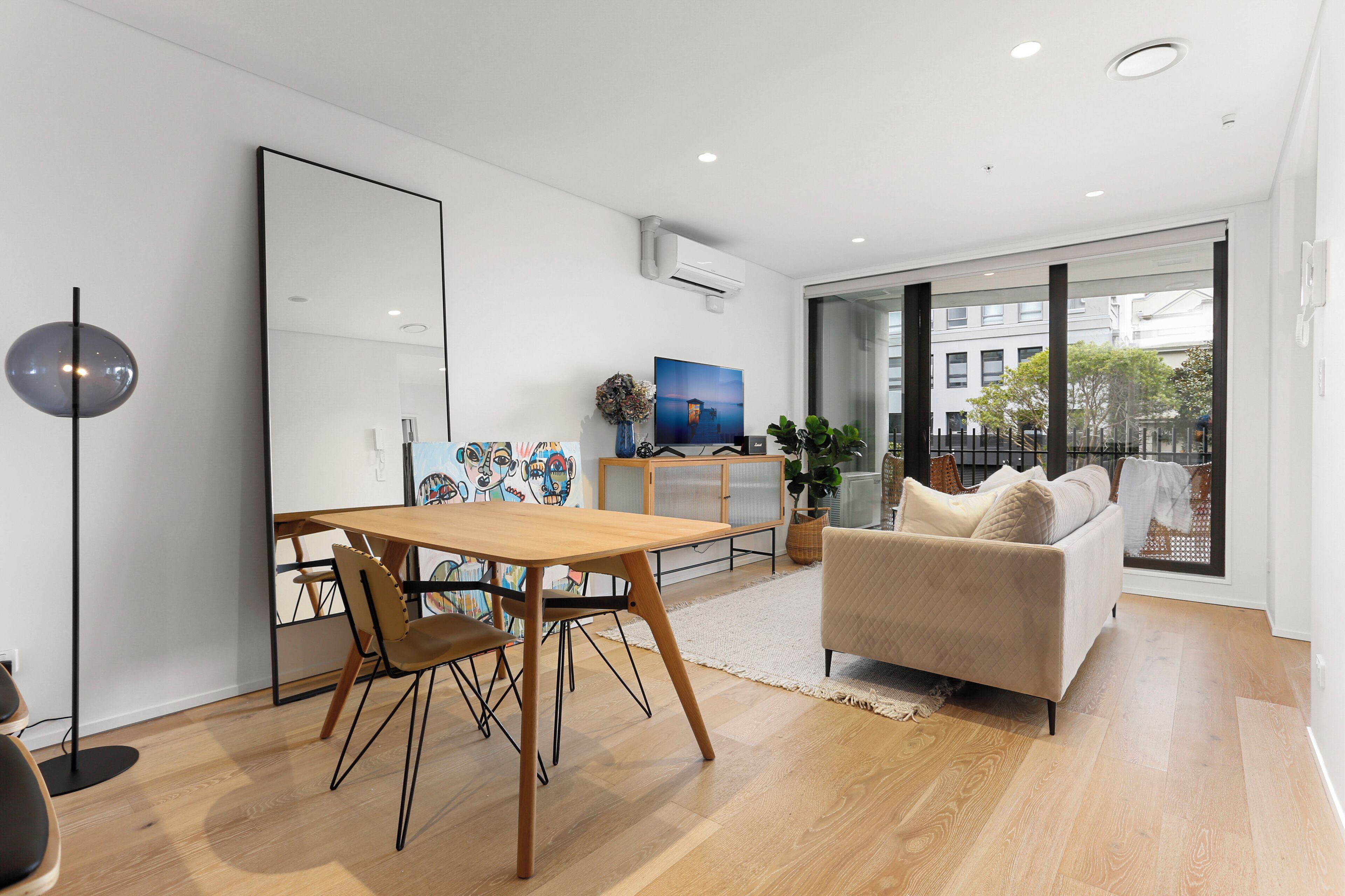 A modern living room with a wooden dining table, a sofa, and a large window offering a view of the outside.