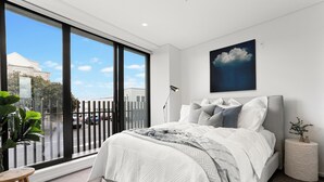 A modern bedroom with a large window, a bed with white bedding, a grey headboard, and a wall-mounted painting of a cloud. - Creative and Carefree Living (Auckland)