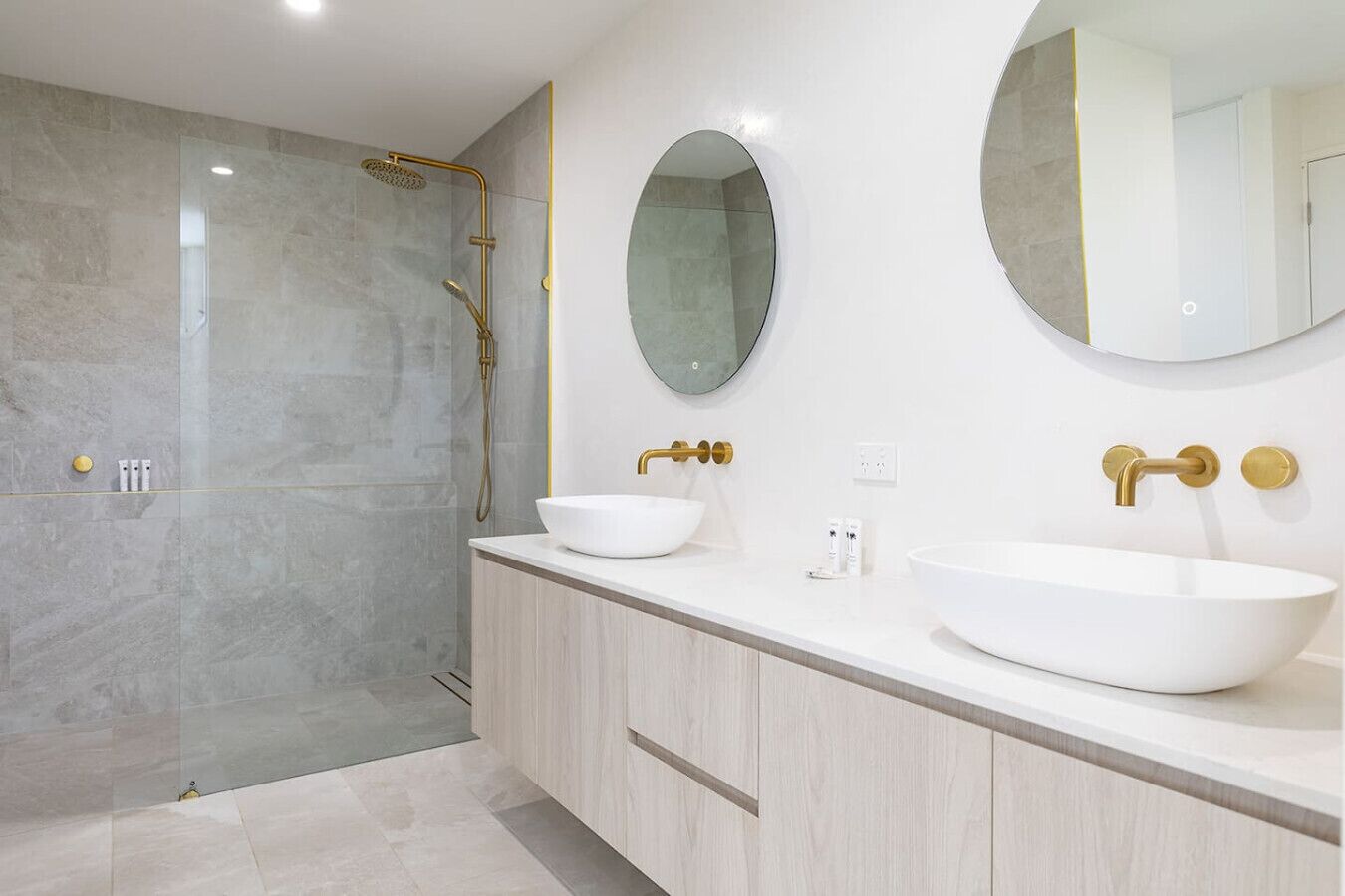 A modern bathroom with a glass-enclosed shower, two white vessel sinks, and gold fixtures.