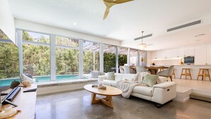 Ein geräumiger Wohnbereich mit großem Fenster zum Pool, ein weißes Sofa, ein Couchtisch und eine Küchenzeile mit Holzstühlen.