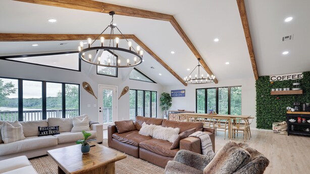 Un salon spacieux avec un grand canapé en cuir marron, un divan blanc et un fauteuil à fourrure. On y trouve une table basse en bois, une table à manger en bois avec des chaises, et une grande fenêtre offrant une vue sur l’extérieur.