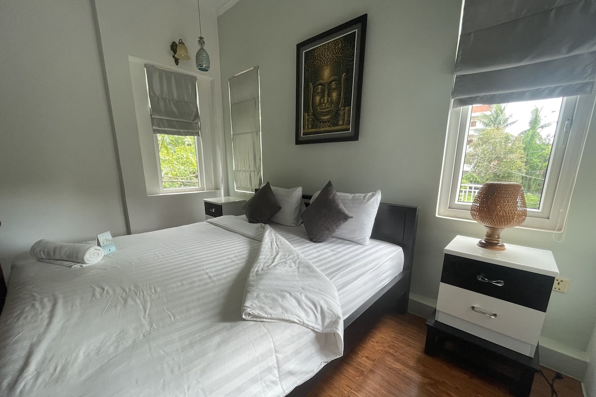 A neatly made bed with pillows, a bedside table with a lamp, and a window with a view of greenery.