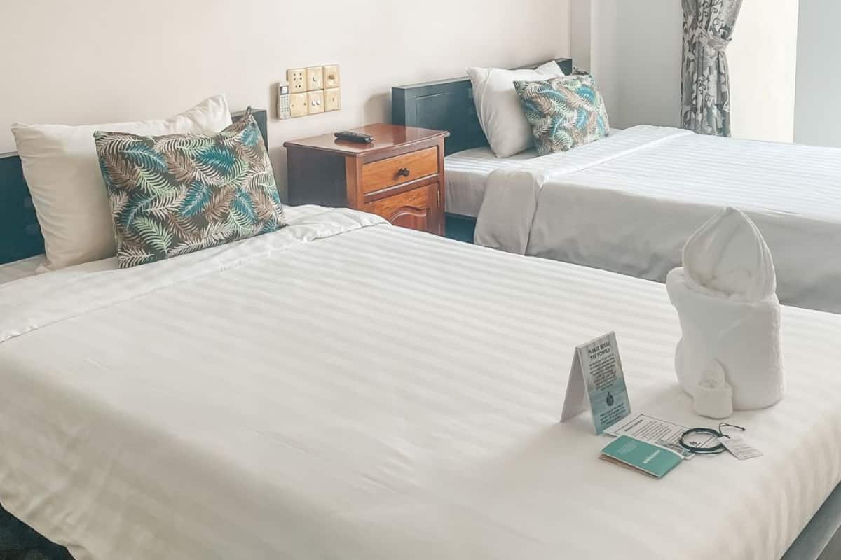 A hotel room with two beds, each with white linens and decorative pillows. There is a wooden nightstand with a remote control and a brochure on it.