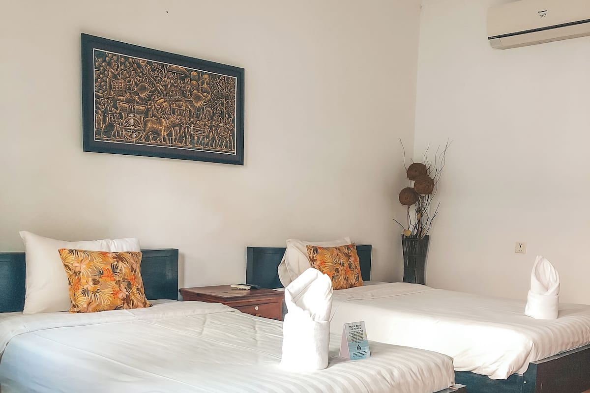 A hotel room with two beds, a wooden floor, a framed artwork on the wall, and a vase with dried flowers.