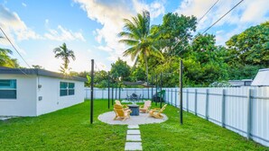 A backyard with a white house, a swing set, and a seating area with chairs and a table. - Miami Backyard Oasis • 4BR • Pool Table • Pergola • Spacious Outdoor Retreat (North Miami Beach)