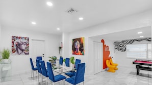 A modern dining area with a glass table and blue chairs, a pool table, and colorful artwork on the walls. - Miami Backyard Oasis • 4BR • Pool Table • Pergola • Spacious Outdoor Retreat (North Miami Beach)