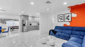 A modern living room with a blue sofa, a glass coffee table, and a kitchen in the background. - Miami Backyard Oasis • 4BR • Pool Table • Pergola • Spacious Outdoor Retreat (North Miami Beach)