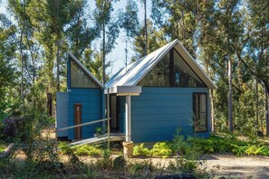 A small, modern cabin with a steeply pitched roof and large windows, surrounded by dense forest. - The Bower At Broulee (Broulee)