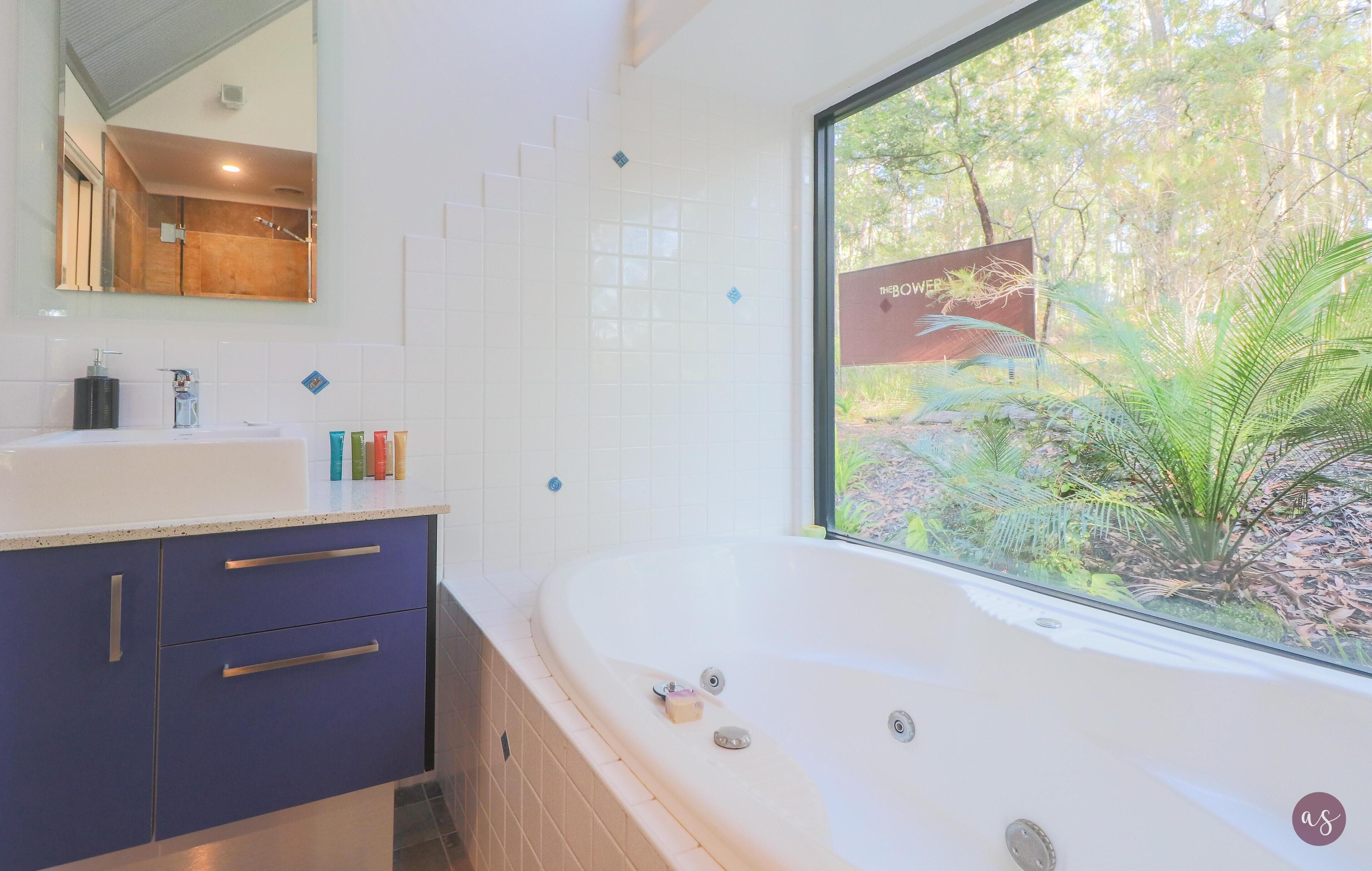 A modern bathroom with a large bathtub, a sink with toiletries, and a window with a view of greenery.
