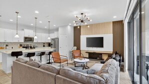 Ein modernes Wohnzimmer mit einer großen Couch, einem Couchtisch und einem Fernseher. Es gibt eine Küchenzeile mit weißen Schränken sowie einen Essbereich mit Stühlen.