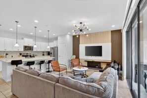 A modern living room with a large sofa, a coffee table, and a TV. There is a kitchen area with white cabinets and a dining space with chairs.