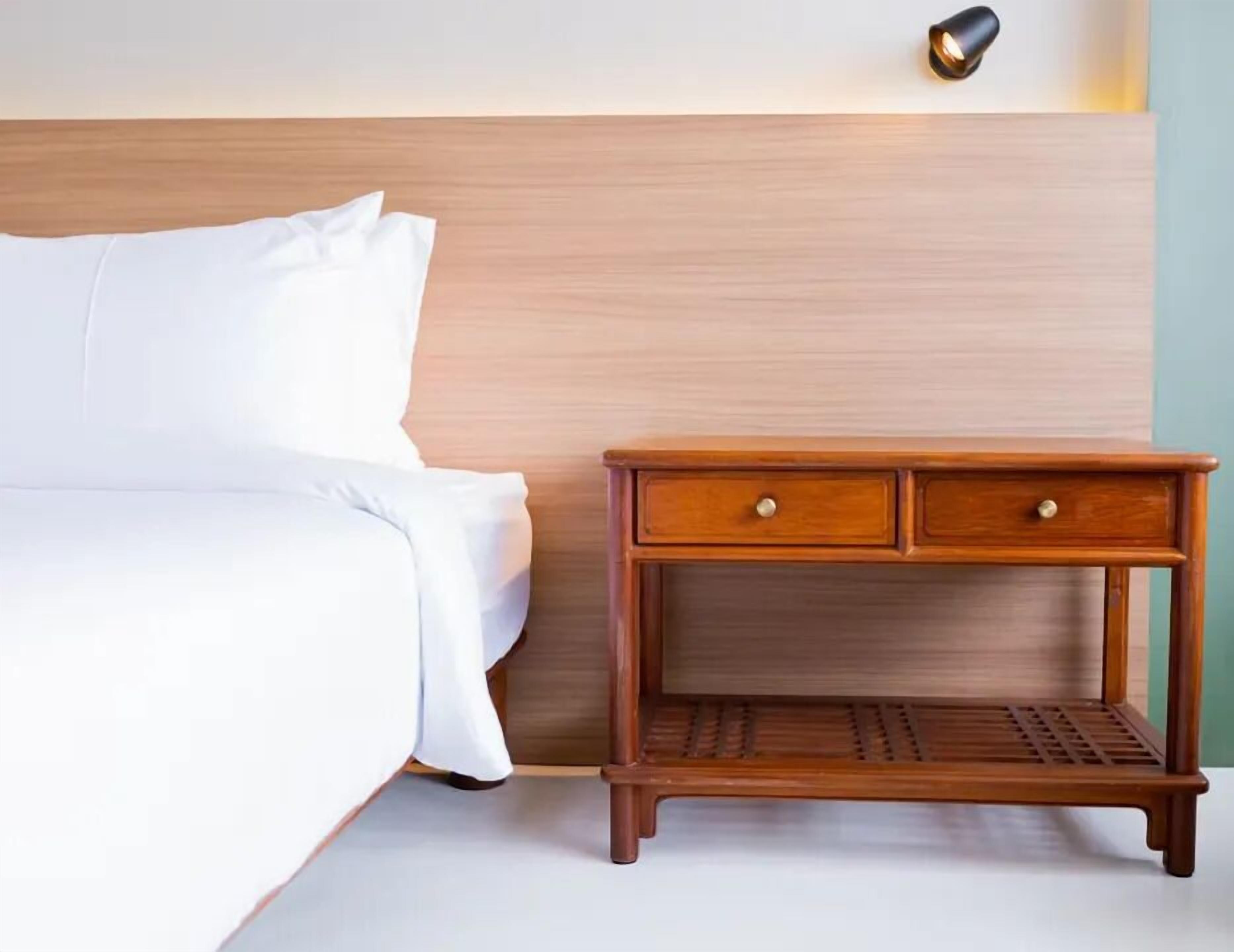 Un lit bien fait, avec du linge de lit blanc, une table de chevet en bois dotée de deux tiroirs et un luminaire fixé au mur.