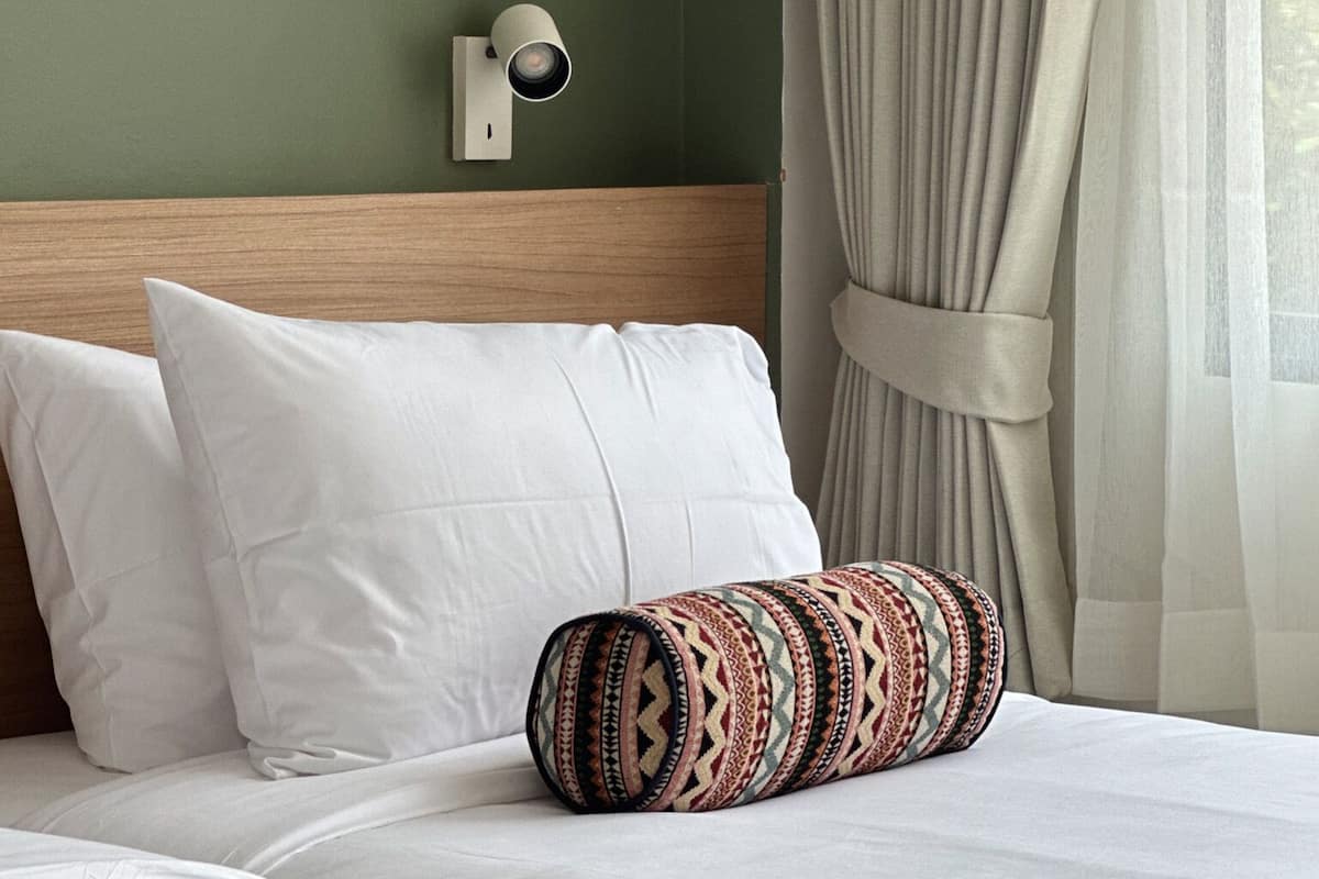 A hotel room with a bed, white pillows, a patterned pillow, a wall-mounted light, and a window with curtains.