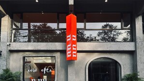 Entrada de hotel con un amplio ventanal que muestra el cartel "Now Open" y un estandarte rojo con letras blancas.