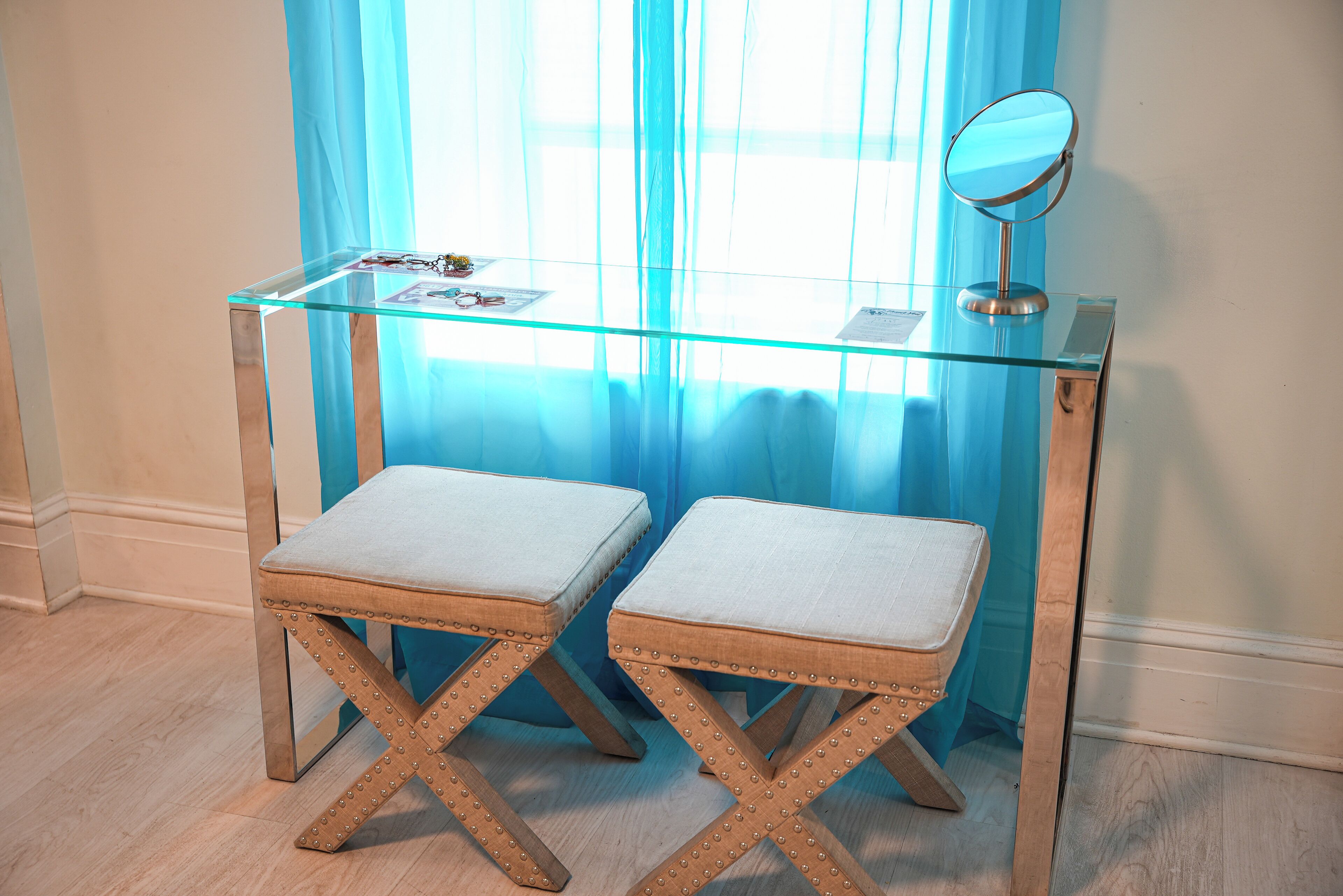 Een glazen kaptafel met een spiegel, twee comfortabele krukken en een klein plankje met spullen erop.