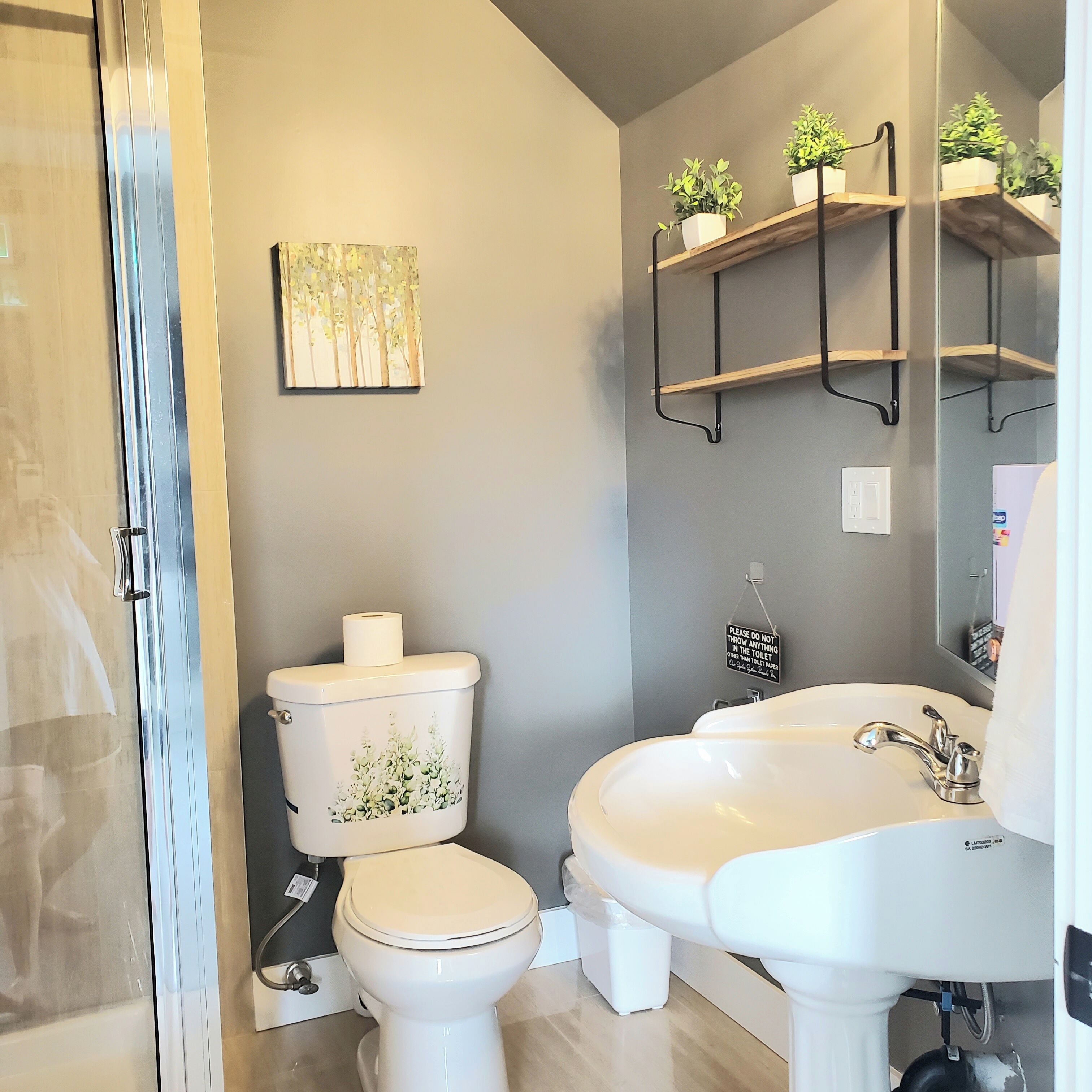 A bathroom with a toilet, sink, and shower. There are plants on a shelf above the sink and a painting on the wall.
