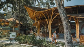 A covered outdoor seating area with bamboo structures, surrounded by tropical plants and trees.