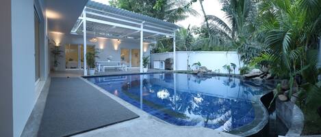 A swimming pool with a covered patio area, surrounded by tropical plants and a white wall.
