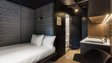 A modern hotel room with a bed, a small desk, a chair, a bathroom with a sink, and a shower.