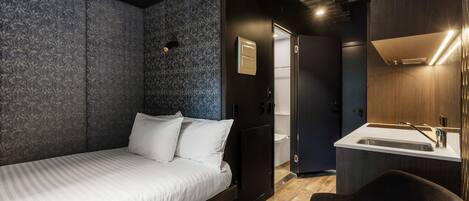 A modern hotel room with a bed, a small desk, a chair, a bathroom with a sink, and a shower.