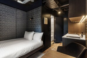 A modern hotel room with a bed, a small desk, a chair, a bathroom with a sink, and a shower.