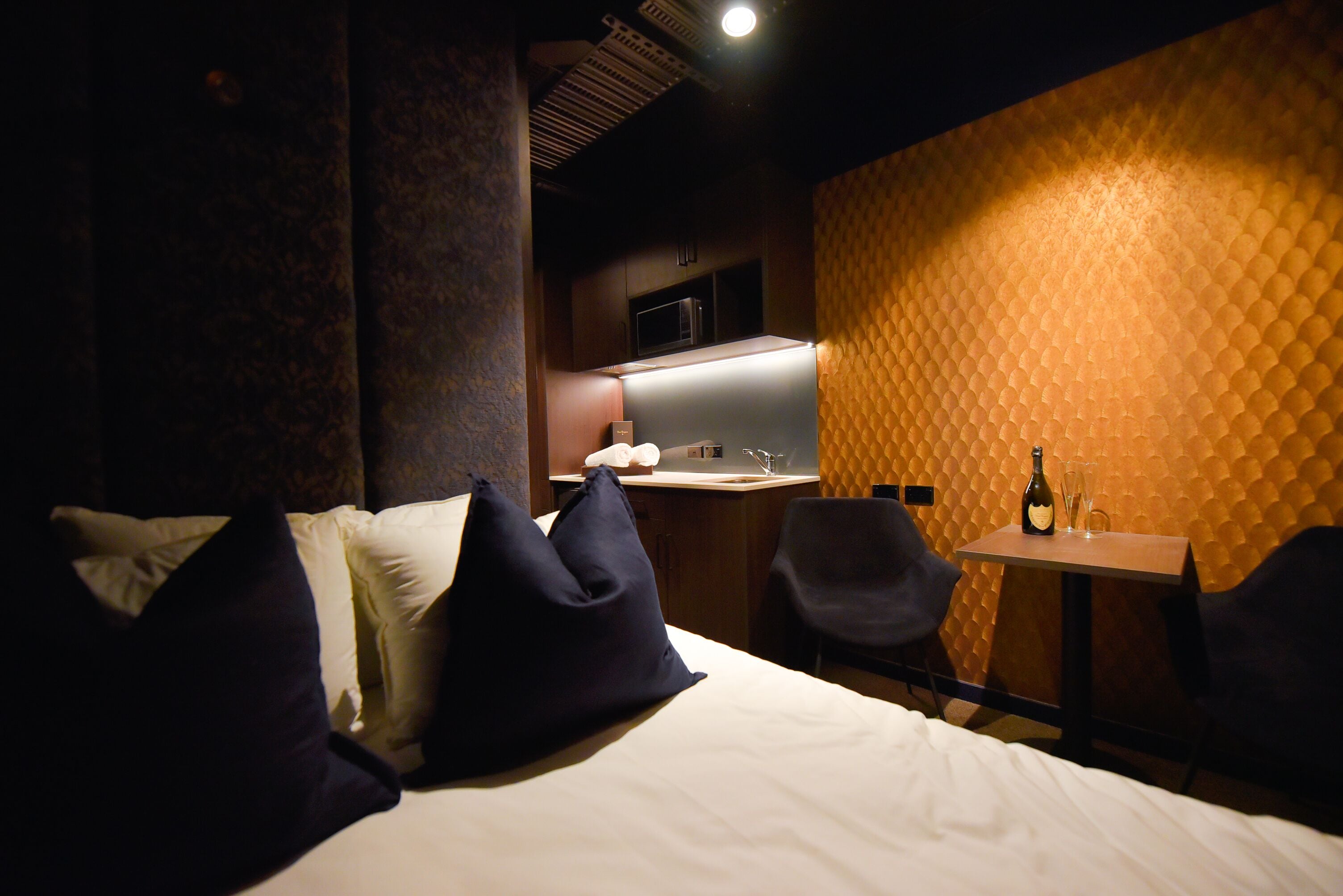 A hotel room with a bed, two chairs, a small table with a bottle and glasses, and a kitchenette in the background.