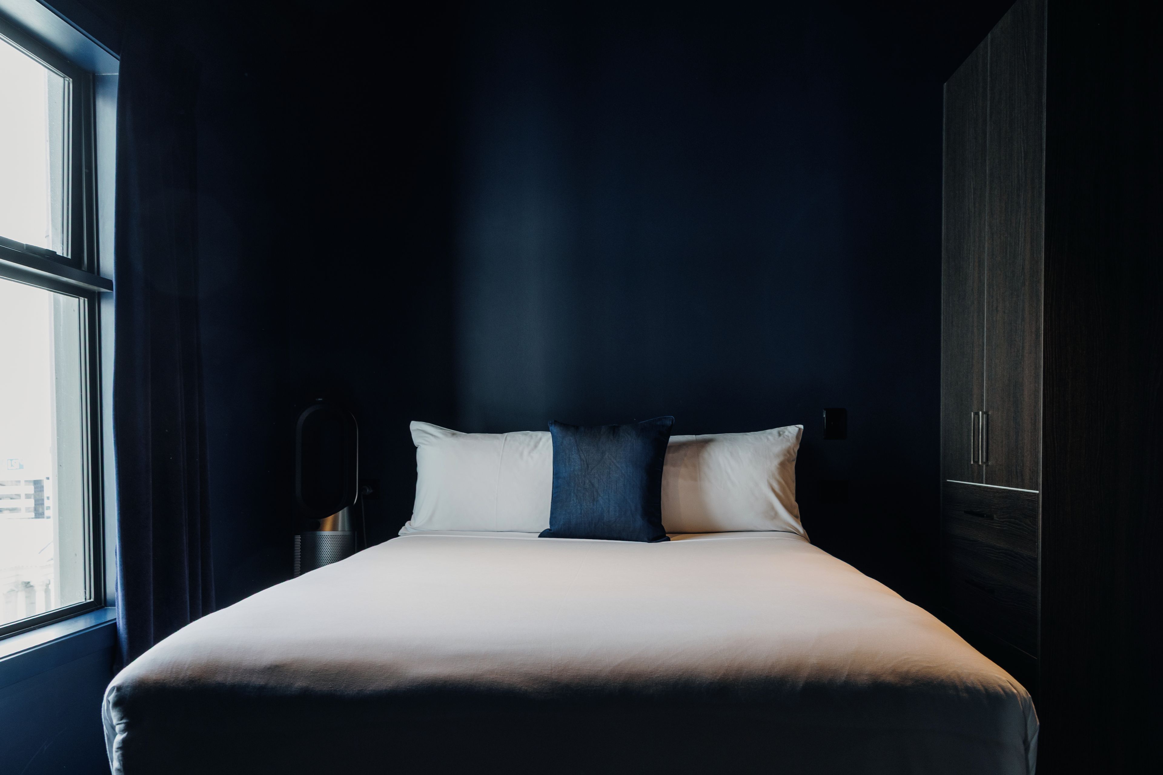 A neatly made bed with a white mattress and two pillows, a window to the left, and a dark wall.