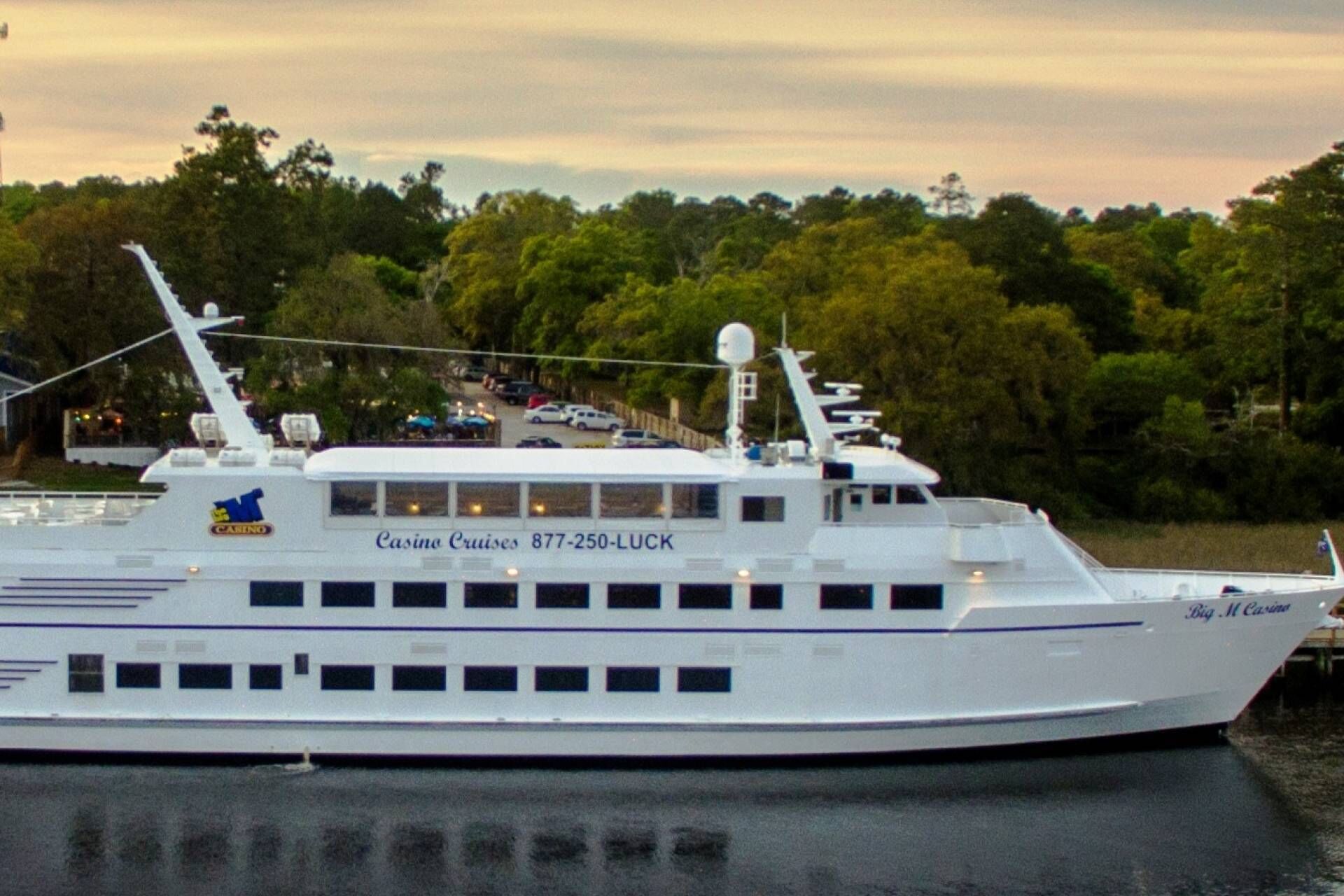 Un gran crucero blanco con varias cubiertas y ventanas, atracado en un puerto con árboles al fondo y un cielo al atardecer.