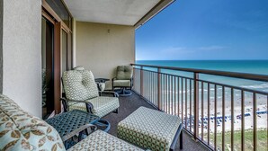 A balcony with two patterned chairs and a table, overlooking a beach with umbrellas.