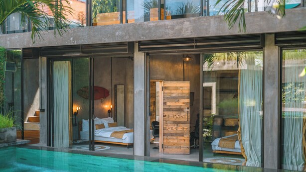 A modern hotel room with a glass wall overlooking a swimming pool, a wooden accent wall, and a cozy bed with a red umbrella. - Villa Konkrete - Tropical 3BR (Kerobokan)