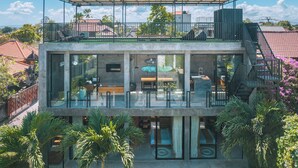A modern two-story house with a glass facade, outdoor seating area, and a covered balcony. - Villa Konkrete - Tropical 3BR (Kerobokan)