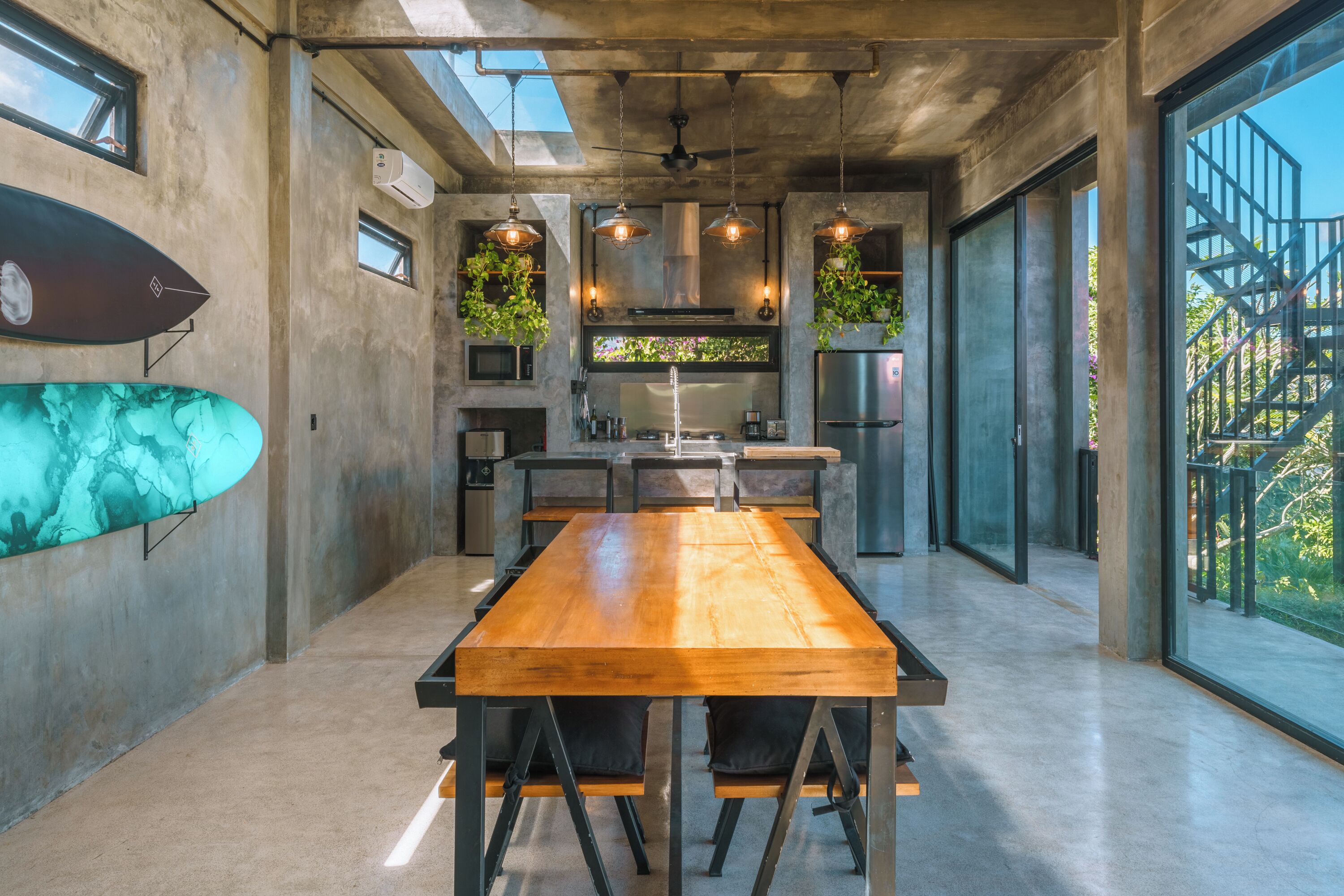 A modern kitchen with a long wooden table, hanging plants, and large windows.
