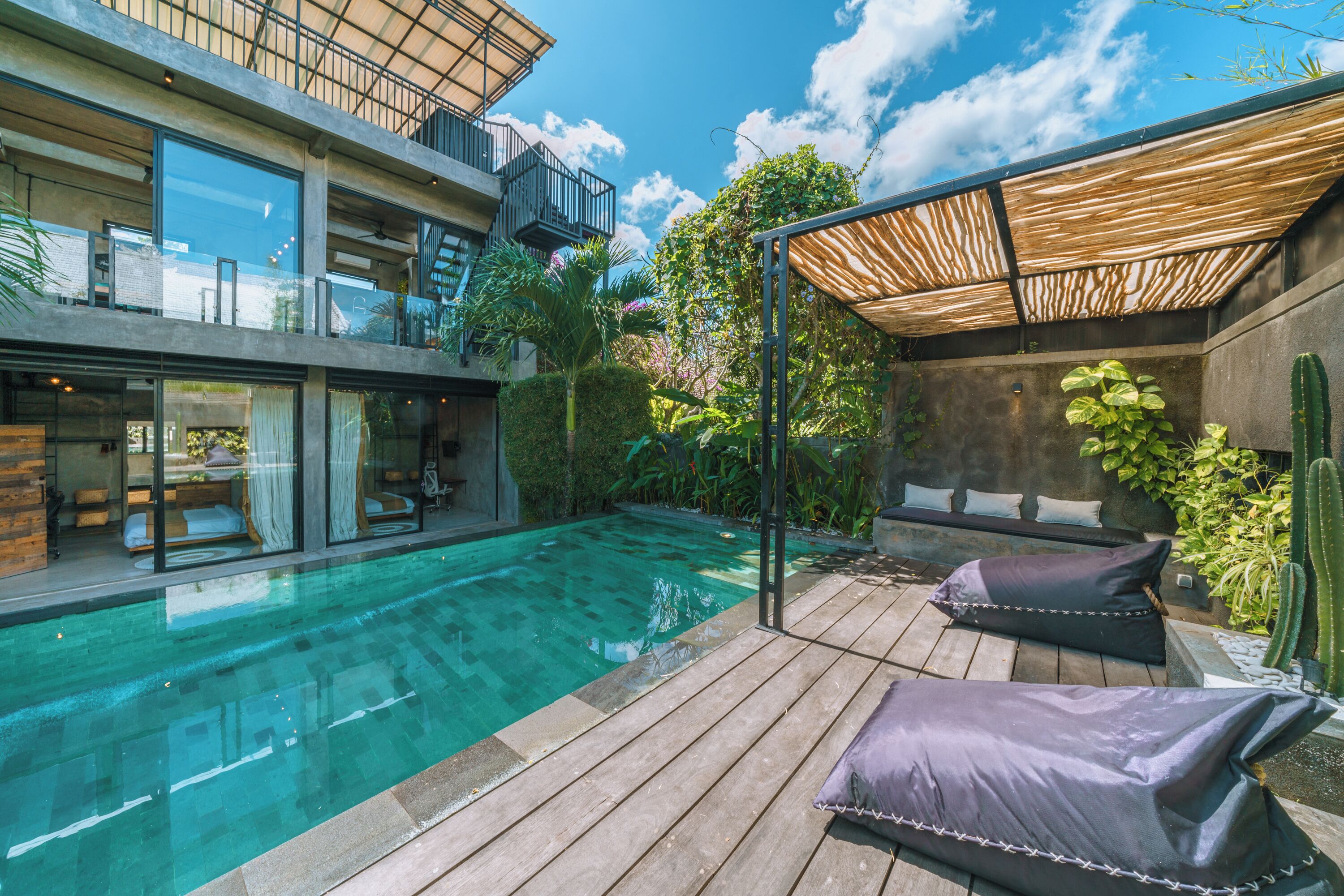 A modern outdoor pool area with a wooden deck, two cushioned seating areas, and a glass-enclosed room with a bed.