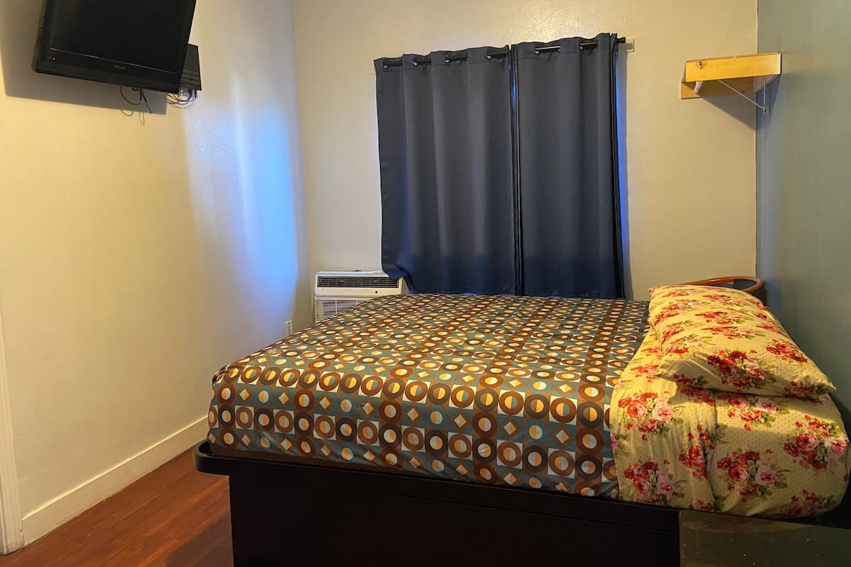 Un dormitorio pequeño con cama, televisión montada en la pared y ventana con cortinas.