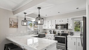 A modern kitchen with white cabinetry, a large island, stainless steel appliances, and pendant lighting. - The Hill House Mtn Views Hot Tub Fire Pit & BBQs (Reno)