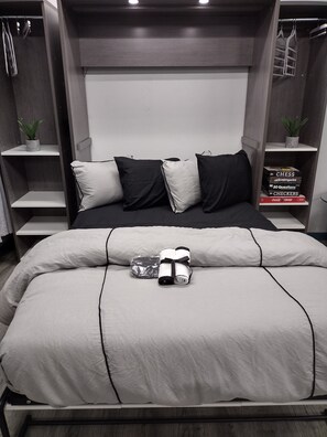 A neatly made bed with a white and black color scheme, a small bedside table with a plant, and a bookshelf with books and a plant. - Luxury In A Quaint Little Package (Oshkosh)