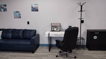 Una habitación con un sofá azul oscuro, una silla de escritorio negra, una mesita con una cafetera y un perchero.