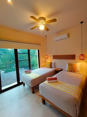 Une chambre d’hôtel avec deux lits, un ventilateur de plafond, un appareil de climatisation, une fenêtre donnant sur un espace verdoyant et une petite table avec une lampe.