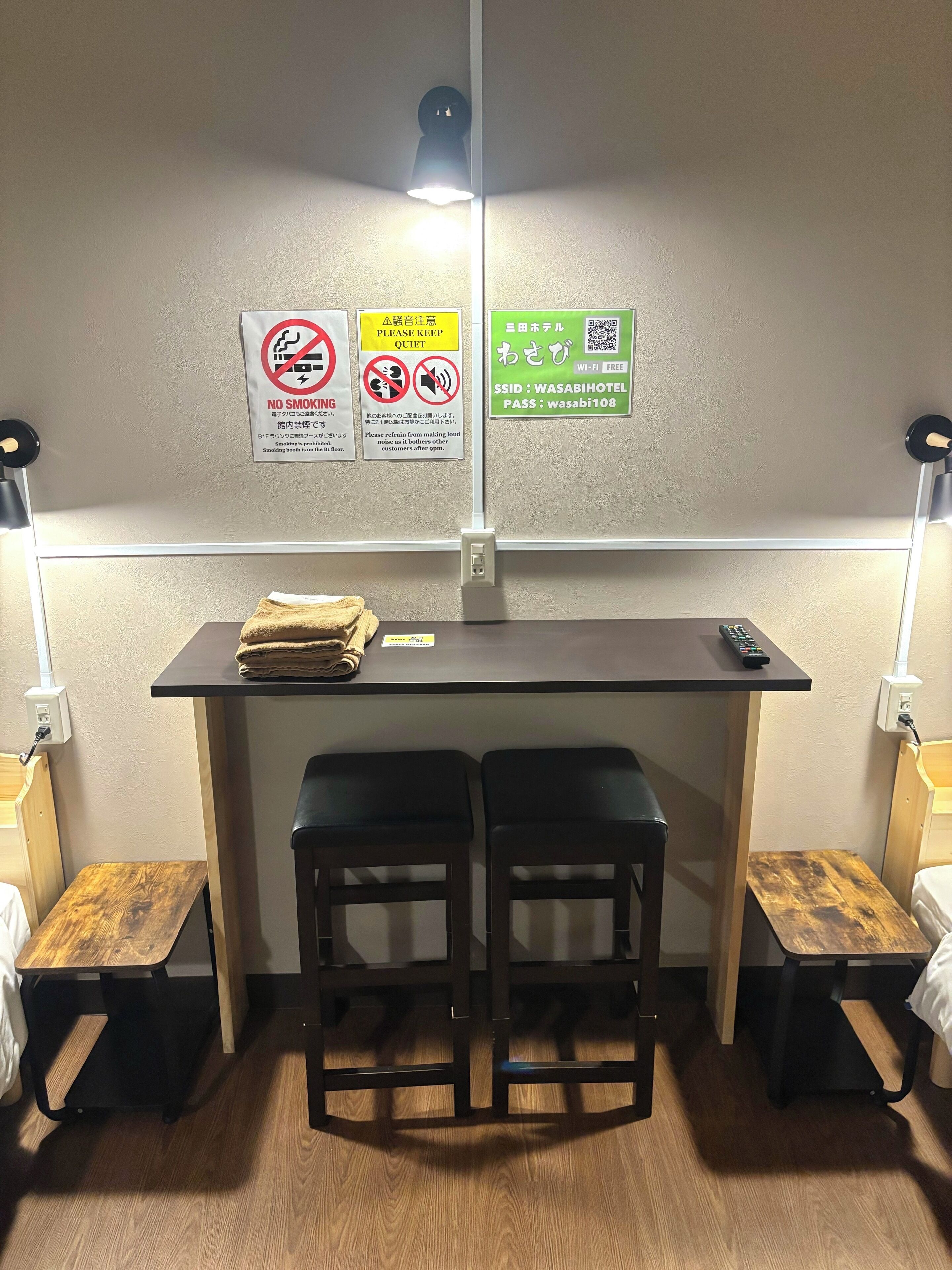 A small hotel room with a desk, two stools, a bed, and a nightstand. There are signs on the wall indicating no smoking and quiet hours.
