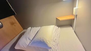 Una cama con ropa de cama y almohadas blancas, un cabecero de madera, una mesita de noche con una lámpara y un estante fijado a la pared.