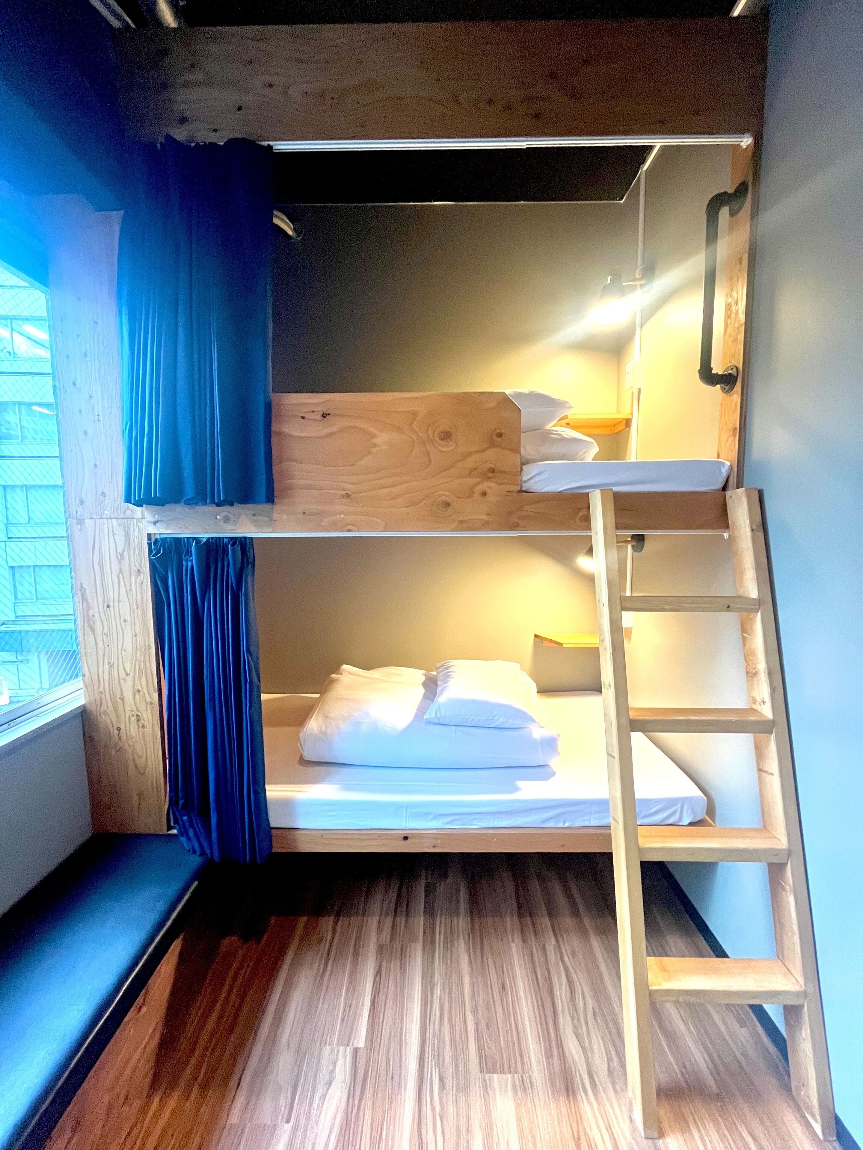 A bunk bed with a wooden ladder, a window with blue curtains, and a wooden floor.