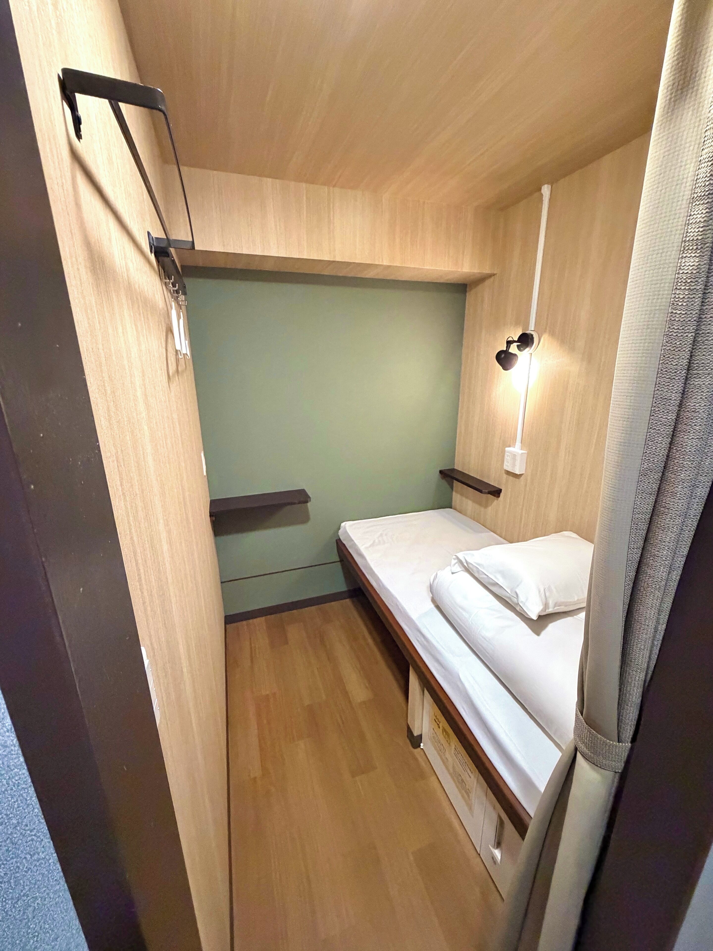 A compact room with a single bed, a small shelf, a wall-mounted light, and a curtain.